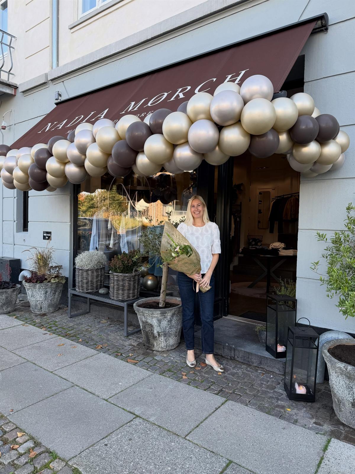 Nadia Mørch med en buket blomster foran butikken på Strandvejen, pyntet med balloner